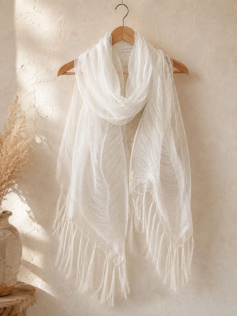 May include: A lightweight, semi-sheer white scarf with a delicate, crinkled texture and long, flowing fringe along the bottom edges. The scarf is draped over a wooden hanger, showcasing its soft, airy fabric and elongated rectangular shape.