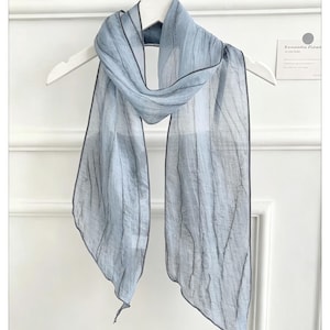 Könnte beinhalten: Ein hellblauer Schal mit einer transparenten, knittrigen Textur und dunkelgrauen Kanten. Der Schal hängt über einem weißen Kleiderbügel und zeigt eine lange, rechteckige Form mit vertikalen Falten im gesamten leichten, halbtransparenten Stoff.