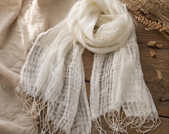 May include: A lightweight, off-white scarf featuring a loose, semi-sheer woven texture. The scarf has a soft, crinkled appearance with long, delicate fringe detailing along both ends, creating a flowing and airy look for this accessory.