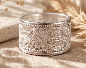 May include: A silver ring with an intricate filigree design. The ring features a wide band with detailed scrollwork and beaded edges. The openwork pattern allows light to pass through, creating a delicate and elegant appearance. The ring is likely made of sterling silver.