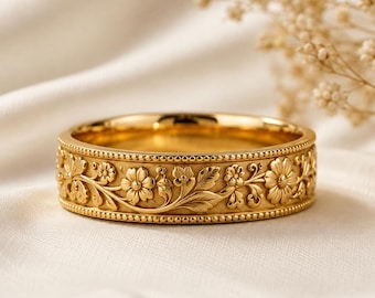 May include: A gold band ring featuring an intricate embossed floral pattern with vines and leaves. The ring has a matte finish and is bordered by delicate beaded edges along the top and bottom, creating a textured and detailed metallic surface.