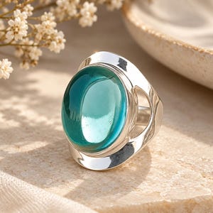 Può includere: Un anello in argento con una grande pietra ovale traslucida color verde acqua incastonata in una fascia di metallo lucido. La fascia presenta un design ampio e aperto che si curva attorno alla gemma centrale, creando una superficie liscia e riflettente.