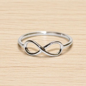 May include: A silver ring with an infinity symbol design. The ring is displayed on a light wooden surface, highlighting its polished finish and simple elegance. A delicate piece of jewellery.