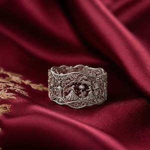 May include: A wide silver ring featuring intricate filigree metalwork. The band displays a detailed scene with two figures in long dresses standing among trees and a small structure, all rendered in a delicate, openwork silver design.