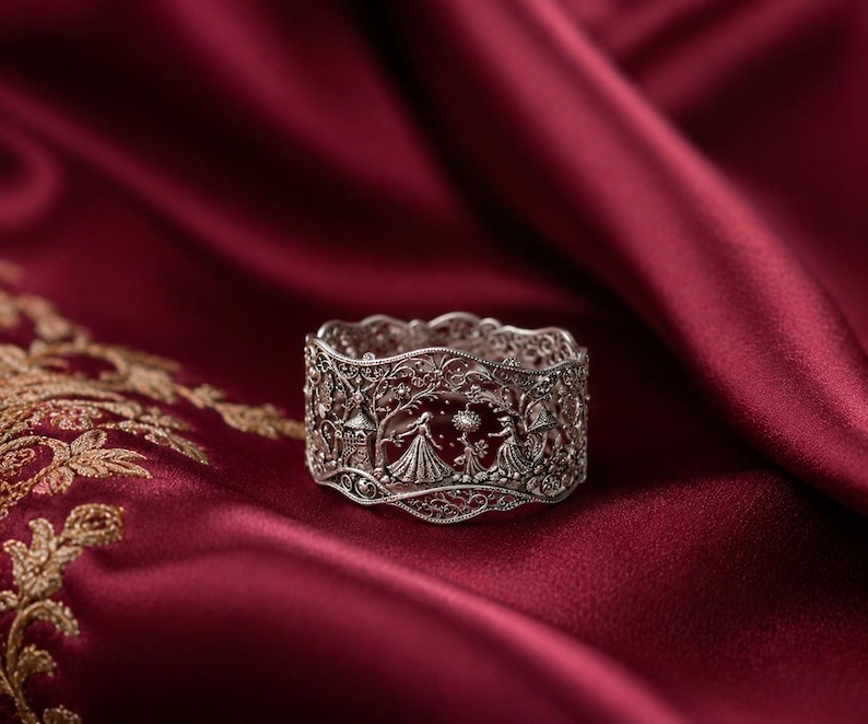May include: A wide silver ring featuring intricate filigree metalwork. The band displays a detailed scene with two figures in long dresses standing among trees and a small structure, all rendered in a delicate, openwork silver design.