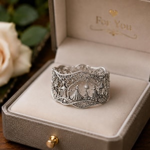 May include: A silver-toned filigree ring featuring an intricate relief design of figures in gowns, trees, and castle-like structures. The ring is displayed inside a beige-lined, metallic-colored box with the text For You printed inside the lid.