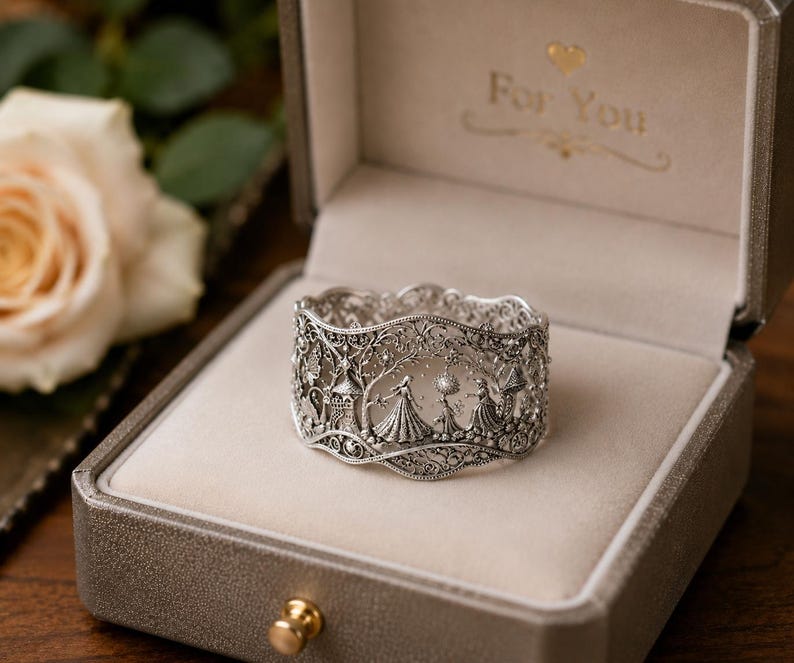 May include: A silver-toned filigree ring featuring an intricate relief design of figures in gowns, trees, and castle-like structures. The ring is displayed inside a beige-lined, metallic-colored box with the text For You printed inside the lid.