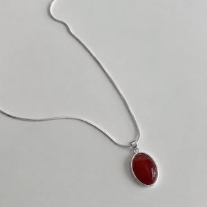 Könnte beinhalten: Eine silberne Halskette mit einem roten, ovalen Anhänger. Der Anhänger ist in einer silbernen Lünette gefasst und hängt an einer feinen Silberkette. Die Halskette wird auf weißem Hintergrund präsentiert.