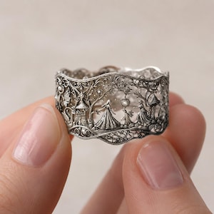 May include: A wide silver band ring featuring intricate filigree metalwork. The surface displays a detailed relief scene of figures in gowns, small structures, and foliage, with a scalloped top and bottom edge and an oxidized finish.