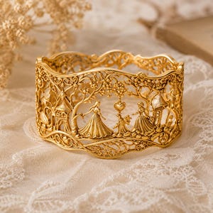 May include: A gold-toned metal cuff bracelet featuring an intricate openwork design. The surface displays a relief scene with two figures in long dresses, a small animal, a heart shape, and architectural structures surrounded by foliage.