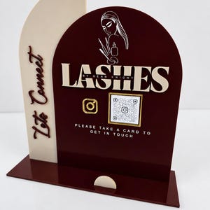 May include: A burgundy and cream acrylic sign featuring the text LASHES BY GENN KNIGHT, Let's Connect, and PLEASE TAKE A CARD TO GET IN TOUCH. The sign includes a white line art graphic of a person, an Instagram icon, and a square QR code.