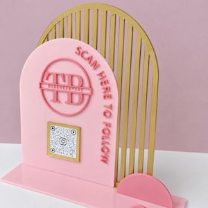 May include: A pink acrylic sign with a gold arched frame. The sign features a circular logo with the text TD ESSENCEBYTAY, a square QR code, and the text SCAN HERE TO FOLLOW. It includes a pink base with two small semi-circular accents.