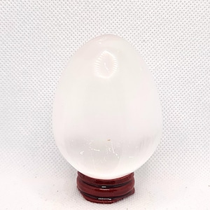 May include: A smooth, translucent white crystal egg with a polished surface, resting securely on a small, circular, dark reddish-brown wooden stand. The egg features subtle internal inclusions and reflects light across its rounded, symmetrical form.