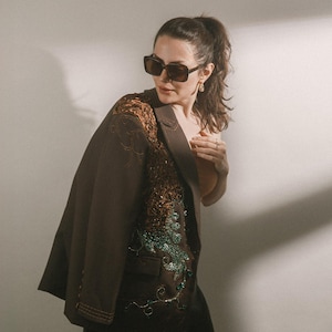 Puede incluir: Un blazer marrón oscuro con intrincados bordados en tonos cobre y adornos de lentejuelas verdes brillantes en el panel frontal. El blazer se lleva sobre una prenda superior de color claro y se combina con gafas de sol oscuras y pendientes dorados.