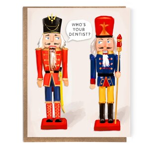 May include: Greeting card featuring two illustrated nutcracker soldiers. One wears a red coat and the other a blue coat. A speech bubble above the blue-coated soldier reads, "WHO'S YOUR DENTIST?"