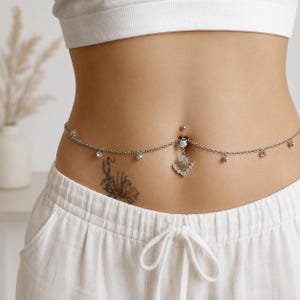 Könnte beinhalten: Eine silberfarbene Bauchketten-Körperkette mit einem Bauchnabelpiercing, das einen fächerförmigen Anhänger mit klaren Kristallen aufweist. Die Kette verläuft um die Taille und ist mit kleinen, gleichmäßig verteilten Kristallakzenten besetzt.