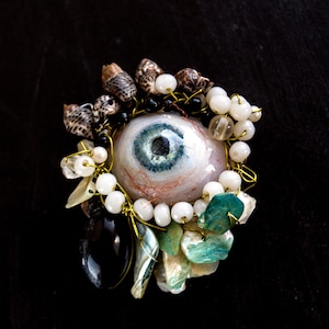 Puede incluir: Un broche que presenta un ojo azul realista central rodeado por un grupo de pequeñas conchas marinas marrones con patrones, cuentas blancas esféricas, fragmentos de conchas verdes iridiscentes y una sola piedra ovalada negra lisa, todo unido con alambre.