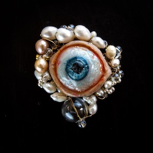 Könnte beinhalten: Eine Brosche mit einem zentralen, realistischen blauen Auge, umgeben von einer Anordnung aus weißen und pfirsichfarbenen Perlen, klaren Glasperlen und dünnem goldfarbenem Draht. Das Auge hat eine detaillierte Iris und Pupille in einem fleischfarbenen Rahmen.