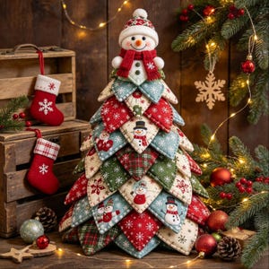 Op de afbeelding: Een decoratief kerstboomornament opgebouwd uit gelaagde, driehoekige stoffen lapjes in rood, groen en crème, voorzien van sneeuwpop- en sneeuwvlokpatronen. Een pluche sneeuwpopfiguur met een rode sjaal zit bovenop de boomstructuur.