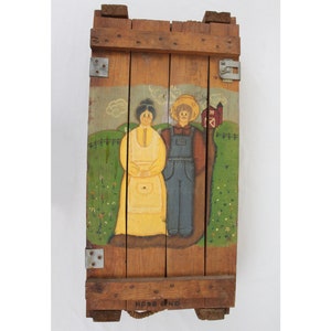 May include: A wooden crate painted with a scene of a couple standing in a field with a red barn in the background. The crate has a rope handle and the words "NEARING" printed on the bottom.