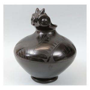 May include: A dark brown ceramic vessel with a rounded body and a detailed, stylized owl-like head. The vessel features etched geometric patterns and a small, circular base. The surface has a glossy finish.