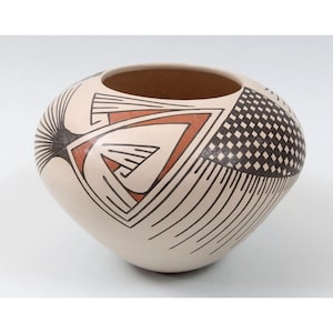 May include: A cream-colored ceramic bowl with a wide opening. The bowl features geometric designs in black and reddish-brown, including a sunburst, a checkered pattern, and angular shapes. The bowl has a rounded shape and a smooth surface.
