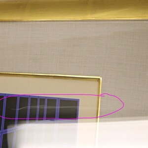 May include: A framed artwork with a gold-colored frame. The artwork features a geometric design with blue and black rectangular shapes. The background is a light beige fabric.