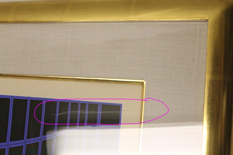 May include: A framed artwork with a gold-colored frame. The artwork features a geometric design with blue and black rectangular shapes. The background is a light beige fabric.
