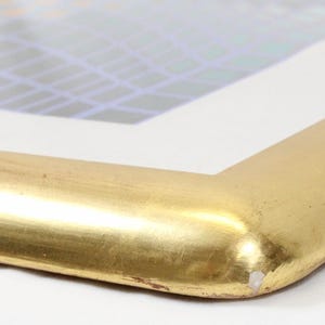 May include: Close-up of a gold-colored picture frame corner. The frame has a rounded edge and a shiny, metallic finish. The artwork inside the frame has a geometric design with blue and white colors.