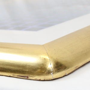 May include: Close-up of a corner of a gold-colored picture frame. The frame has a rounded edge and a metallic finish. The corner is joined with a mitered cut. The frame is set against a white background.