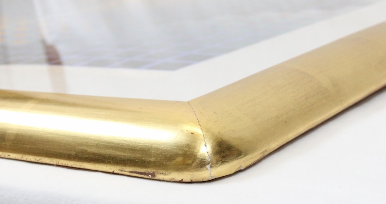 May include: Close-up of a corner of a gold-colored picture frame. The frame has a rounded edge and a metallic finish. The corner is joined with a mitered cut. The frame is set against a white background.