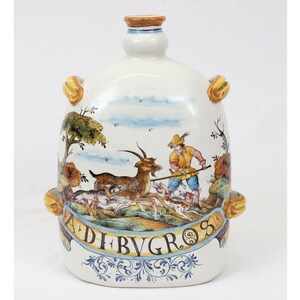May include: A white ceramic bottle with a hunting scene painted on it. The scene depicts a man with a staff chasing a goat and two dogs. The bottle has a gold trim and the words "ADI BVGROS" written on it.