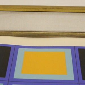 May include: Abstract art print featuring a repeating pattern of colorful squares. The design includes black, yellow, and light blue squares set against a purple background. The print is framed with a gold-colored frame.