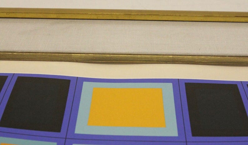 May include: Abstract art print featuring a repeating pattern of colorful squares. The design includes black, yellow, and light blue squares set against a purple background. The print is framed with a gold-colored frame.