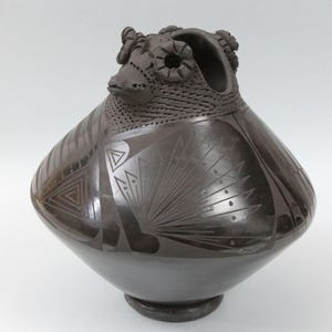 May include: A dark brown ceramic vessel with a rounded base and a detailed ram's head design at the top. The vessel features etched geometric patterns and a textured neck, showcasing traditional craftsmanship.