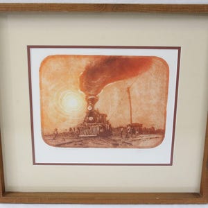 May include: Framed sepia-toned print of a steam train emerging from a bright sun. The train is centered, with smoke billowing upwards. The print is set within a cream-colored mat and a wooden frame.