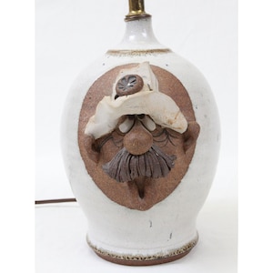 May include: A ceramic lamp with a white base and a brown, circular face design. The face features a mustache, beard, and hat. The lamp has a brass-colored top and a brown electrical cord.