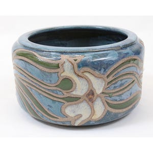 May include: A round ceramic bowl featuring a glazed finish in shades of blue and green. The exterior is decorated with a raised, light-colored floral pattern that contrasts with the darker blue background, creating a textured surface design.