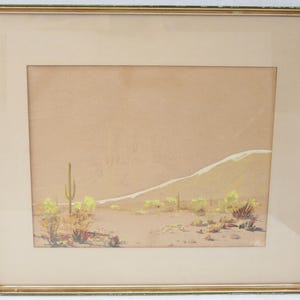May include: Framed desert landscape art featuring a saguaro cactus and other desert plants. The artwork is in a gold-toned frame with a cream-colored mat. The painting depicts a desert scene with a mountain in the background.