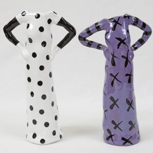 May include: Two ceramic sculptures of women in dresses. One dress is white with black polka dots, the other is purple with black X patterns.