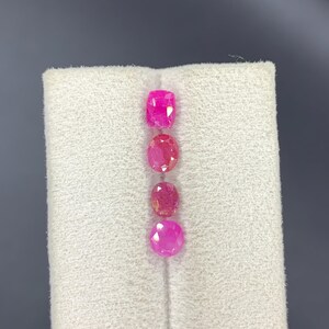 May include: Four loose faceted gemstones arranged in a vertical line. The stones are vibrant pink in color and feature various cuts, including cushion, oval, and round shapes, displaying clear internal facets and reflective surfaces.