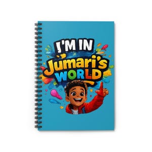 Puede incluir: Un cuaderno de espiral con una cubierta azul claro. La cubierta presenta el texto "I'M IN Jumari's WORLD" y una ilustración de dibujos animados de un niño sonriente que señala hacia arriba, con salpicaduras de pintura coloridas.
