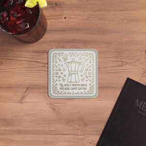 May include: Square coaster with a light gray border and a white center featuring a gold-colored coffee pot design and the Italian text "La vita è troppo breve per bere caffè cattivo."