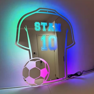 以下が含まれることがあります： サッカーボールのグラフィックが下部にあり、中央にSTANという名前と数字の10がプリントされた、ジャージ型の壁掛けミラー。ミラーは鮮やかなLEDライトストリップで照らされており、縁の周りに青と紫の光を放っています。