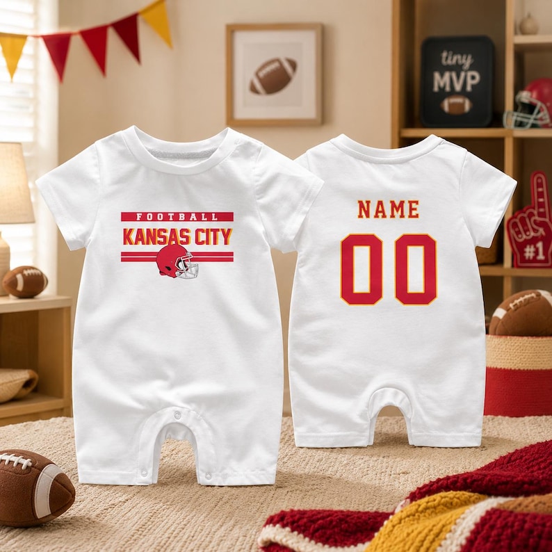 May include: A white short-sleeve baby romper featuring a red and yellow graphic on the front with the text FOOTBALL KANSAS CITY and a football helmet. The back displays the text NAME above the number 00 in large red and yellow lettering.