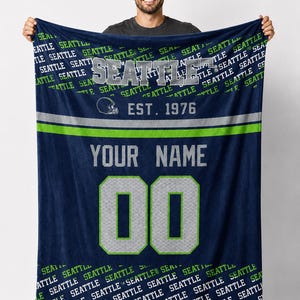 Könnte beinhalten: Marineblaue Fleece-Decke mit dem Wort "SEATTLE" in großen, silbernen Buchstaben. Die Decke zeigt eine Footballhelm-Grafik, den Text "EST. 1976" und Platz für einen Namen und eine Nummer. Das Design ist in Silber und Grün gehalten.