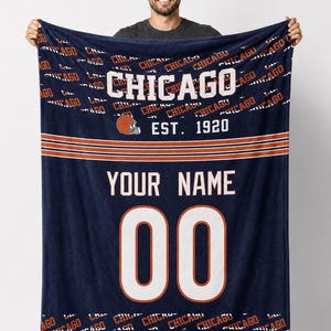 Może przedstawiać: Granatowy koc z napisem "CHICAGO" w białych literach. Koc posiada grafikę kasku do futbolu i napis "EST. 1920". Poniżej znajduje się napis "YOUR NAME" i numer "00" w kolorze białym. Obecne są również pomarańczowe i białe paski.