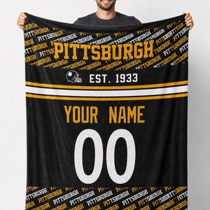 Puede incluir: Manta negra con "PITTSBURGH" en letras doradas, un gráfico de casco de fútbol y "EST. 1933". Cuenta con una franja blanca y dorada, con espacio para "YOUR NAME" y el número "00". La manta está sujeta por una persona.