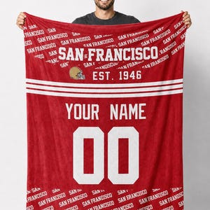 Puede incluir: Manta polar roja con texto "SAN FRANCISCO" y un gráfico de casco de fútbol. Presenta el texto "YOUR NAME" y el número "00". La manta es de color rojo vibrante con letras y rayas blancas.