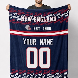 May include: A navy blue blanket with the words "NEW ENGLAND" in white and red. The blanket has a football helmet graphic and the text "EST. 1960". It features a space for a name and the number "00".